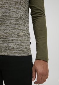 Långärmad skjorta med ett grått texturerat mönster och ärmar i olivgrönt. Tyget är mjukt, sittande, med en etikett på sidan. Svarta byxor synliga.