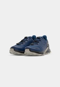 Des chaussures de sport bleues avec une tige texturée, des accents noirs et une semelle extérieure grise. Elles sont dotées d'un système de laçage sécurisé et d'un design de talon rembourré.