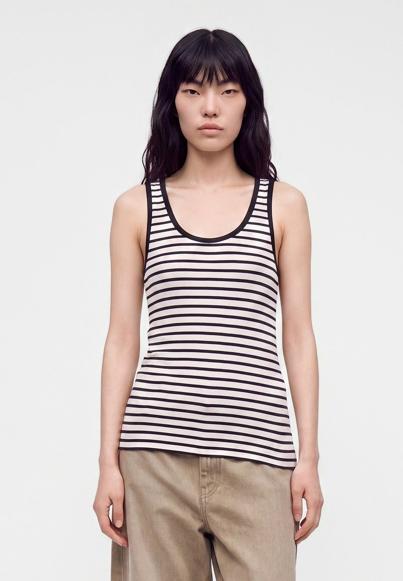 Jeune femme aux cheveux noirs lisses portant un débardeur blanc à rayures horizontales noires et un pantalon beige, debout devant un fond uni.