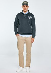 Tumehall pullover krae ja valge kirjaga, hele sinine särk all, beežid püksid ja valged spordijalatsid. Oliivroheline müts.