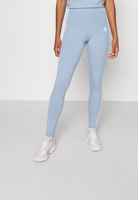 Osoba oblečená v světle modrých legínách Adidas s bílými bočními pruhy a bílých teniskách, stojící na jednobarevné světle šedé podlaze.