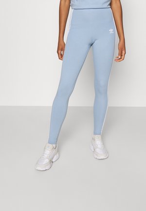 Person wearing light blue Adidas leggings with white side stripes and white sneakers, standing on a plain light grey floor.