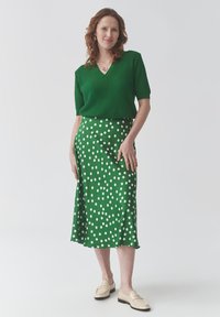 Haut en tricot vert à manches courtes avec un col en V, associé à une jupe midi verte à pois. Des chaussures légères à enfiler complètent la tenue.