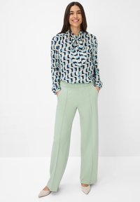 Langärmlige Bluse mit einem geometrischen Muster in Blau, Schwarz und Hellblau; kombiniert mit hochgeschnittenen, mintgrünen, weit geschnittenen Hosen und beigen Schuhen.