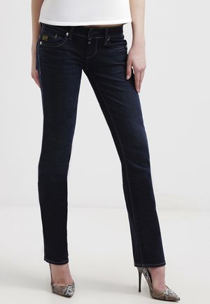 Kobieta w ciemnoniebieskich dopasowanych jeansach i wysokich szpilkach ze skóry węża, stojąca na jednolitym jasnym tle.