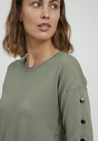 Pull olive avec un col rond, texture côtelée et boutons décoratifs le long des manches. Les coutures ajoutent un subtil intérêt au design.