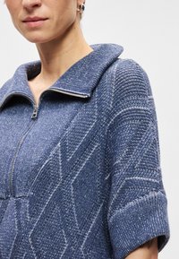 Πλεχτό πουλόβερ σε σκούρο μπλε με κολάρο με φερμουάρ, διακοσμημένο με υφές και κυλιόμενα κοντά μανίκια. Απαλή υφή με ζεστό σχέδιο.