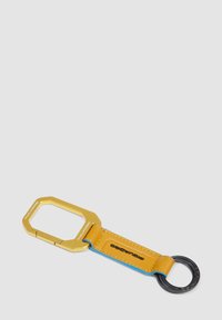 Portachiavi con una cornice ottagonale in metallo dorato e un cinturino in pelle gialla, rifinito con cuciture blu e un anello circolare nero.