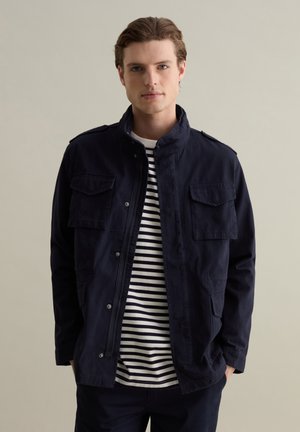 Marineblaue Jacke mit vier Fronttaschen, Stehkragen und Druckknopfverschluss, über einem weiß- und marineblau gestreiften Hemd getragen. Baumwollstoff, lässiges Design.
