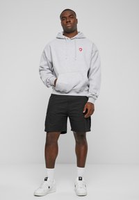 Grauer Hoodie mit rotem Herz-Logo, Fronttasche und Zugband. Kombiniert mit schwarzen Shorts und weißen Sneakers mit schwarzen Akzenten.