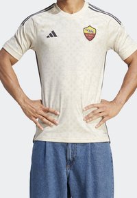 Jersey de Adidas color crema con un sutil patrón de hojas, rayas negras en los hombros y el escudo de AS Roma en el lado izquierdo del pecho. Diseño de manga corta.
