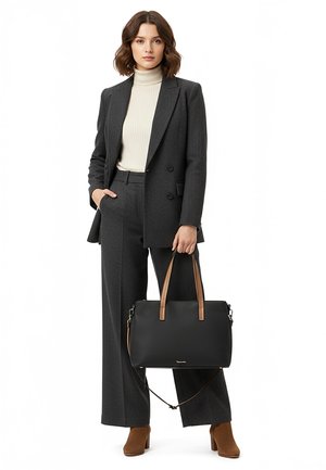 Woman standing wearing dark grey suit with wide-leg trousers, beige turtleneck, brown ankle boots, carrying black handbag with tan straps.