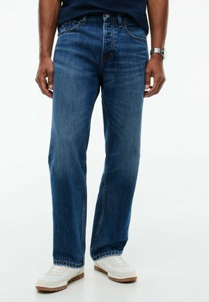 Mand iført blå jeans med lige ben, hvide sneakers og et sølvfarvet armbåndsur mod en ensfarvet baggrund.