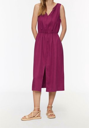 Femme portant une robe midi sans manches violette avec une fente à l'avant et des sandales plates beiges à lanières, se tenant contre un fond blanc.