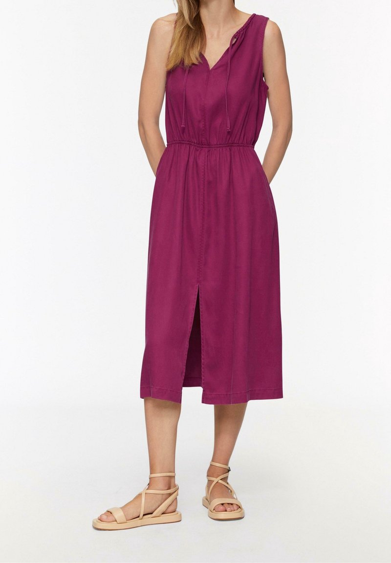 Donna che indossa un abito midi senza maniche di colore viola con spacco frontale e sandali piatti beige con cinturini, in piedi su uno sfondo bianco.