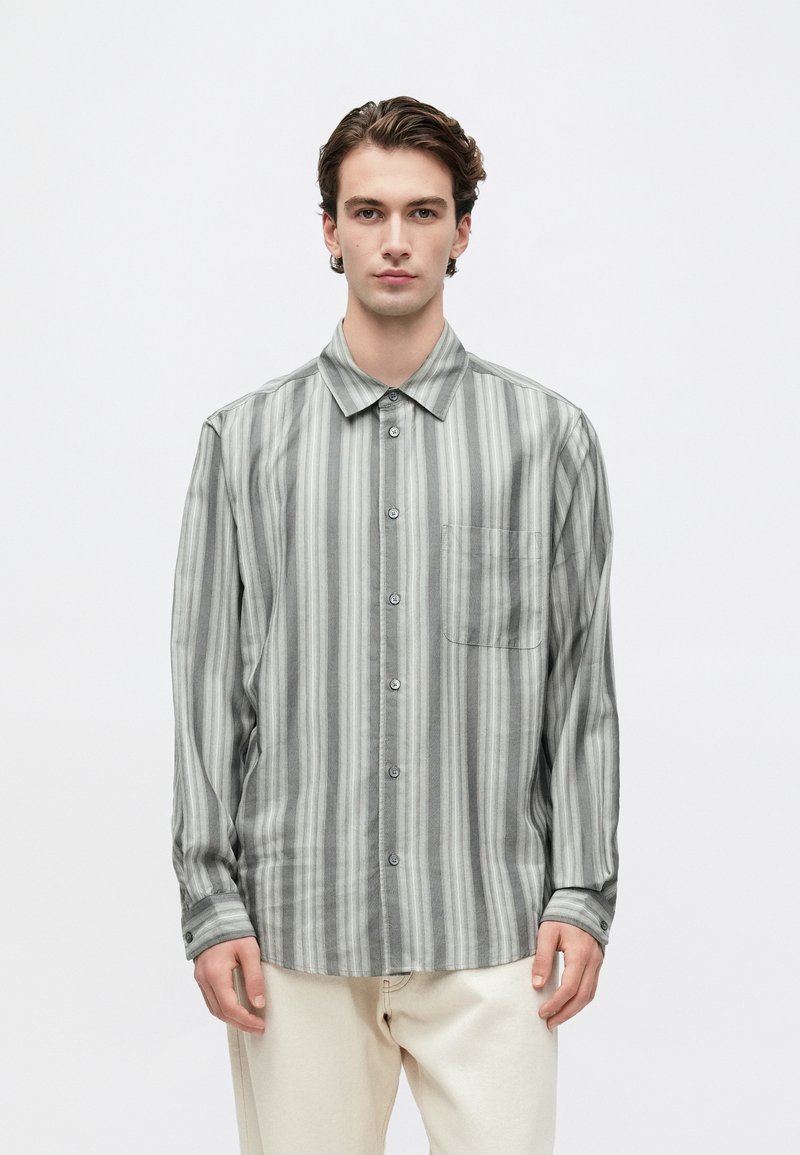 Joven con cabello corto y ondulado que lleva una camisa de botones a rayas gris de manga larga y pantalones ligero color beige, de pie contra un fondo blanco.