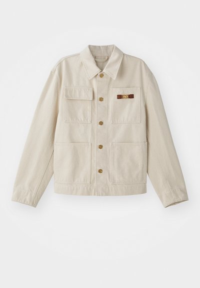 Bež jakna na gumbe z dört sprednjimi žepi, dolgimi rokavi in majhnim rjavim in zlatim okrasnim sponko na desnem žepu na prsih.