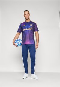Maglietta da calcio con maniche corte in gradiente viola e blu, con accenti gialli e logo "Herbalife", abbinata a pantaloni jogger blu navy e scarpini bianchi.