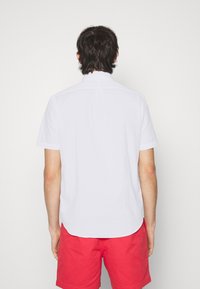 Vit kortärmad skjorta i lätt material, med rak nederkant och subtila sömmar i ryggen, tillsammans med röda shorts.