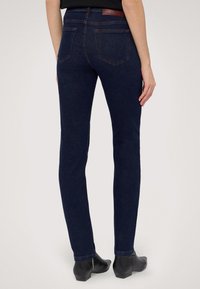 Jean de mezclilla azul oscuro con corte ajustado, que presenta bolsillos traseros y un pequeño parche de cuero con el logo. Textura suave y detalles de costura sutiles.