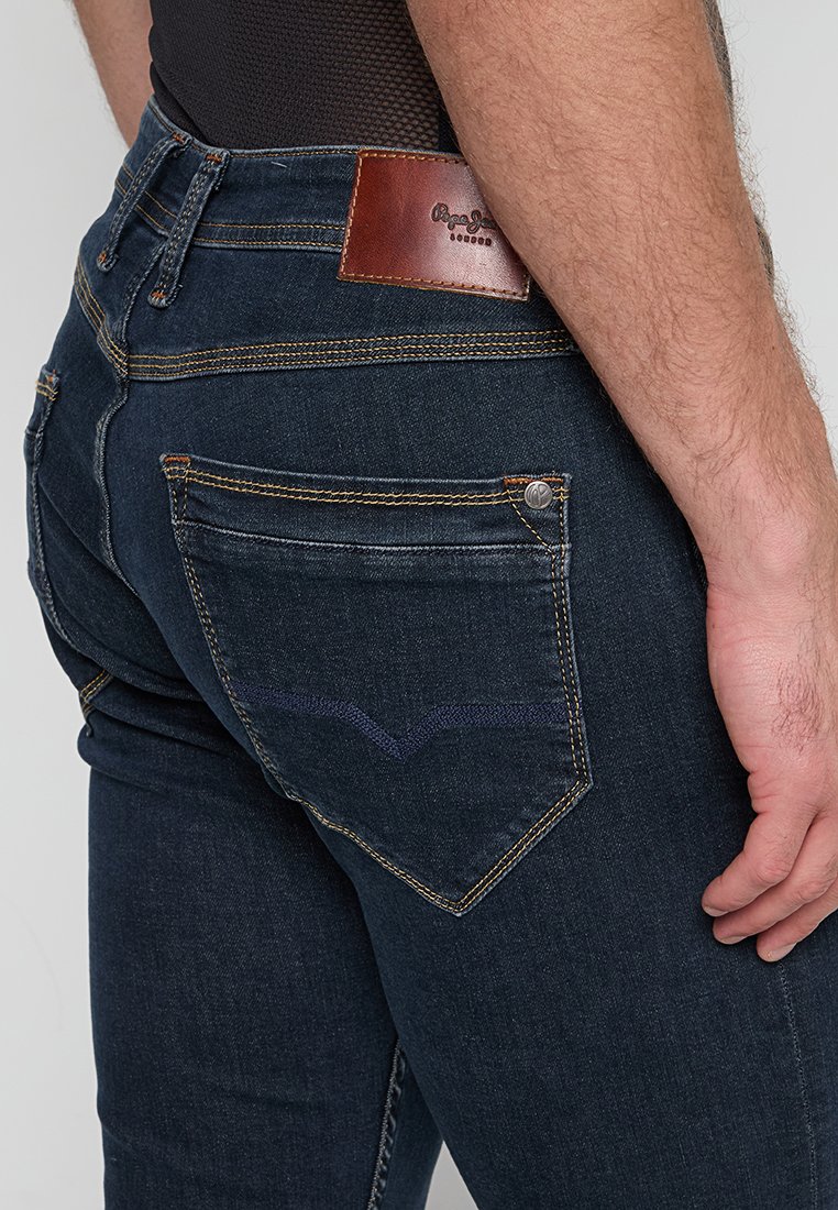 Nahaufnahme des Rückens eines Mannes, der dunkelblaue Jeans trägt, mit einem Leder-Brandlabel und einem auffälligen Taschendesign.