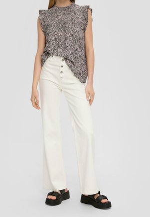 Vrouw draagt een mouwloos patroon top, witte wijde broek met hoge taille en knopen, en zwarte platformsandalen, staand op een grijze achtergrond.