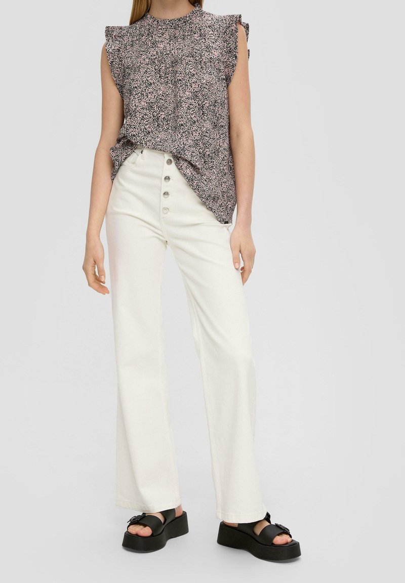 Femme portant un haut sans manches à motifs, un pantalon blanc taille haute à jambes larges avec des boutons, et des sandales plateformes noires, debout sur fond gris.