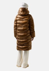 Osoba ubrana w długą metaliczną brązową kurtkę puchową z kapturem, białe spodnie, beżowe buty i białą dzianinową czapkę, stoi tyłem.