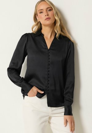 Vrouw met lang blond haar draagt een zwarte satijnen blouse met knopen en een witte broek, leunend tegen een effen lichtgekleurde muur.