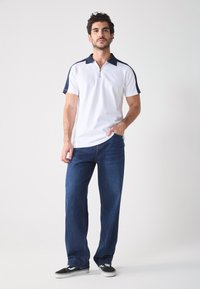Balts zip-up polo ar tumši zilām akcentēm un īsām piedurknēm, kombinēts ar ziliem brīvi pieguļošiem džinsiem un melnām kedām. Minimālistisks dizains.
