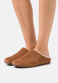 Bruna mockaskor med en slip-on-design, utrustade med en fluffig vit foder och en rund tå. Läderlogotypdetalj på sidan.