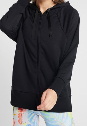 Femme portant un sweat à capuche noir zippé avec cordons de serrage et des leggings à motif feuilles tropicales multicolores, posant avec une main près du visage.