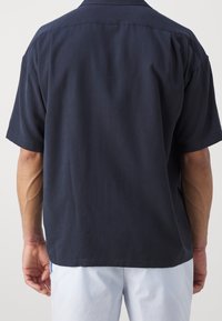 Camisa de manga corta en color marino, hecha de una tela texturizada, con dobladillo recto y cuello clásico. Detalles de costura visibles.