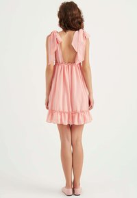 Rosa Kleid mit Rückenschnürung und Schleifen-Trägern, geraffter Taille und vollem Saum. Der Stoff hat kleine Polka-Dots und eine leichte Textur.