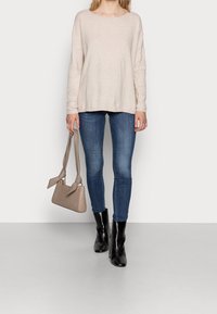 Un haut beige texturé à manches longues, un jean bleu ajusté, des bottines noires à la cheville et un sac à main marron clair avec un détail en nœud.