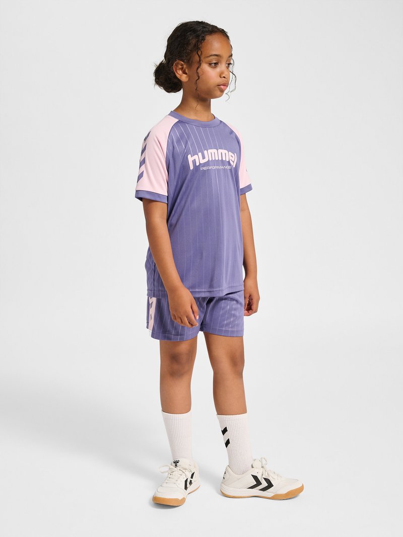 Kind steht seitlich, trägt lila und pinke Hummel-Sportbekleidung mit weißen Socken und weißen Sneakers auf weißem Hintergrund.