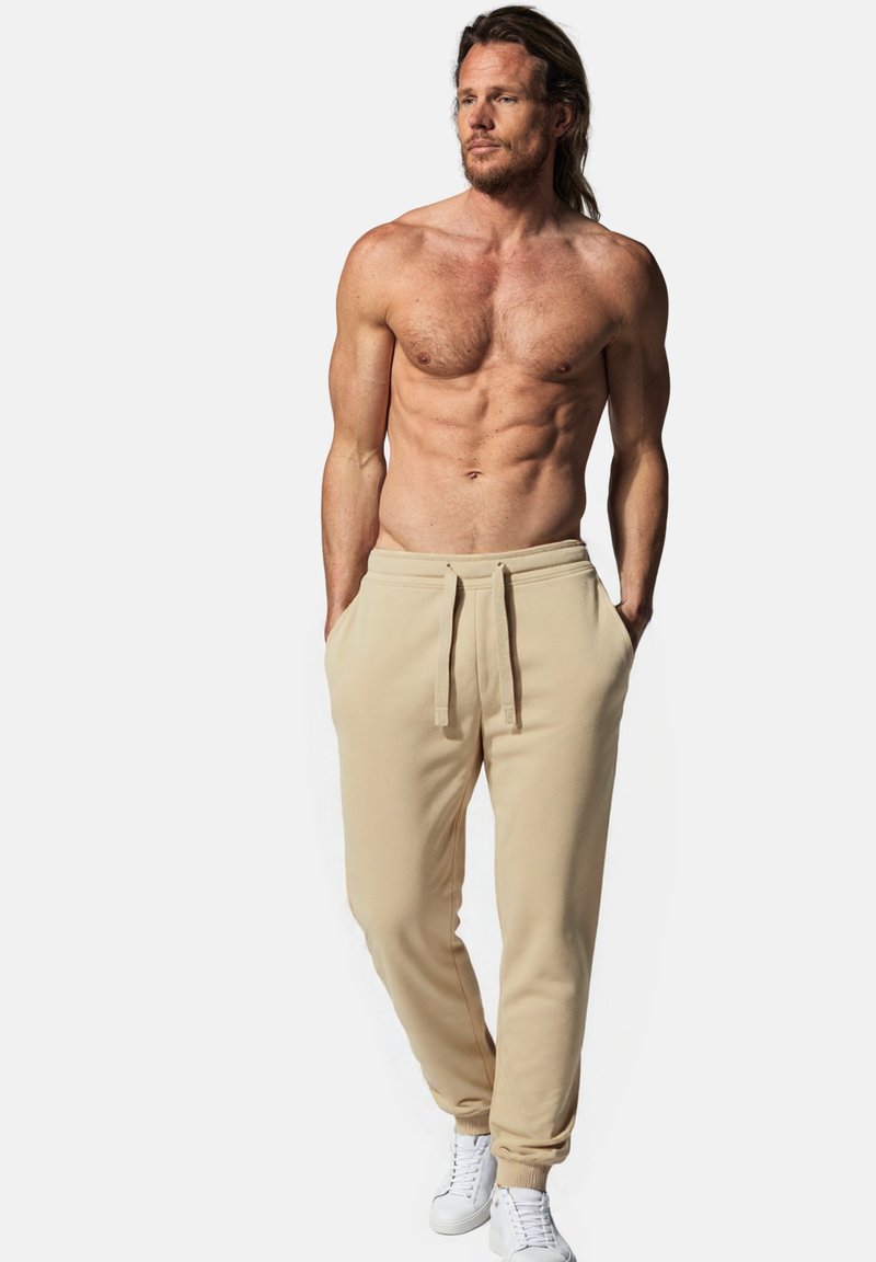 Beige Sweatpants mit elastischem Bund und Kordelzug. Verfügt über seitliche Taschen und gerippte Bündchen. Getragen mit weißen Sneakers.