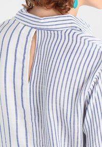Camicia in lino a righe con righe blu e bianche, caratterizzata da un'apertura sul retro e un colletto classico. La texture leggera migliora la traspirabilità.