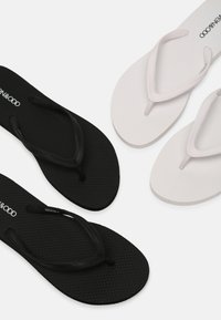 Zwart-witte slippers met gestructureerde zolen en T-bandjes, voorzien van lichte branding op de binnenzolen. De zwarte versie heeft zwarte bandjes, terwijl de witte versie witte bandjes heeft.