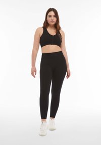 Top sportivo nero con chiusura a zip e leggings neri a vita alta, entrambi realizzati in tessuto elasticizzato, abbinati a sneaker bianche.