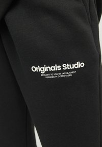 Fekete pulóver, amelyen a "Originals Studio" felirat fehér színben nyomtatva található az ujjon. Az anyag puhanak és texturáltak tűnik.