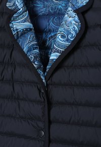 Veste matelassée bleu foncé avec doublure intérieure bleue à motif cachemire et bordure noire le long des bords avant ouverts et de la fermeture par boutons.
