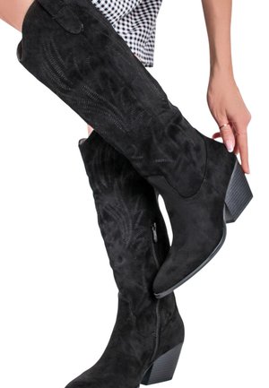 Bottes hautes jusqu'au genou en daim noir avec des motifs brodés et des talons blocs, ajustées sur une personne portant une jupe à carreaux noir et blanc.