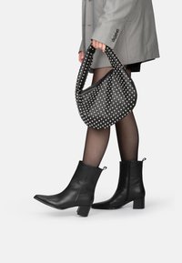 Bottes noires en cuir à cheville avec un bout carré et un talon bloc, associées à un sac à main noir clouté et des collants noirs transparents, le tout sur fond blanc.