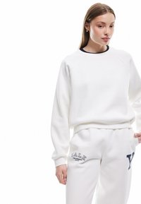 Sudadera blanca con cuello redondo y puños acanalados, combinada con joggers blancos a juego que presentan el texto en navy "YALE UNIVERSITY". Textura de tela suave.