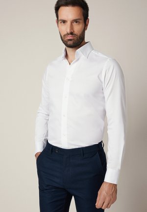 Biała koszula z długim rękawem na guziki, o dopasowanym kroju i klasycznym kołnierzyku, zestawiona z granatowymi spodniami o delikatnej fakturze.
