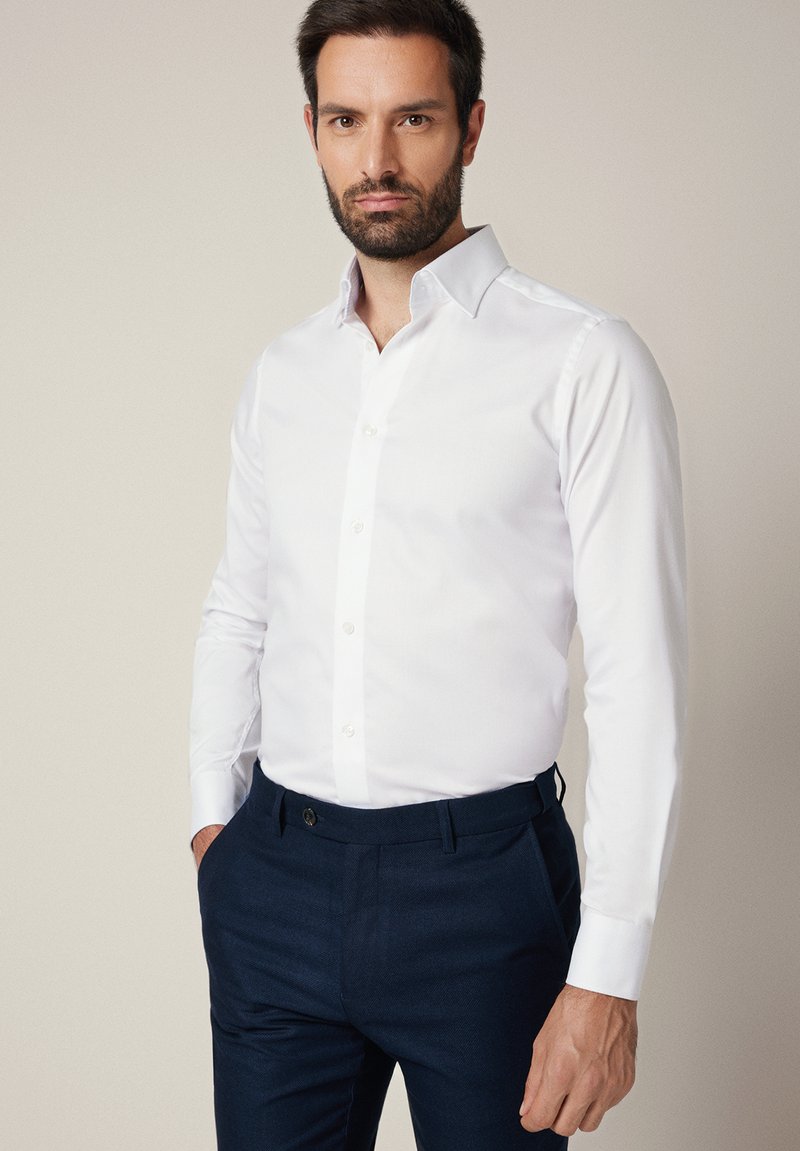 Biała koszula z długim rękawem na guziki, o dopasowanym kroju i klasycznym kołnierzyku, zestawiona z granatowymi spodniami o delikatnej fakturze.