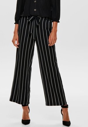 Kobieta ubrana w czarne spodnie z szerokimi nogawkami z białymi pionowymi paskami, z wiązanym w pasie paskiem, w połączeniu z czarnymi szpilkami i czarną bluzką.