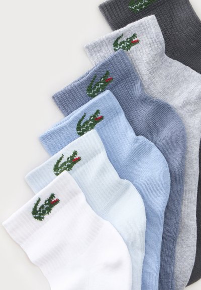 Sept paires de chaussettes pliées en gris, gris clair, bleu, bleu clair et blanc, chacune avec un logo crocodile vert près du bord-côte.