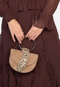 Runde Handtasche aus beigem Wildleder mit einem facettierten Kristallakzent auf der Klappe. Verfügt über einen silbernen, runden Griff und strukturierte Details.